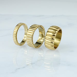 May include: Three gold-toned metal rings with a ridged design. The rings are stacked on a white marble surface.