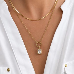 May include: A layered gold-tone necklace set featuring a short chain with small flat circular links and a longer chain with a textured circular ring pendant holding a rectangular clear faceted stone. The jewelry is worn over a white collared shirt.