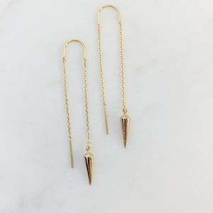 May include: A pair of gold dangle earrings with a pointed, teardrop-shaped charm at the end of each chain.