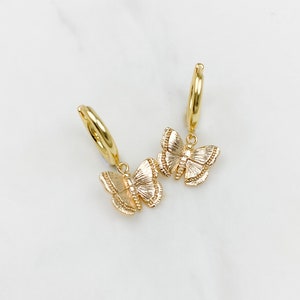 May include: A pair of gold hoop earrings with a dangling butterfly charm. The butterfly charm is textured and has a detailed design.