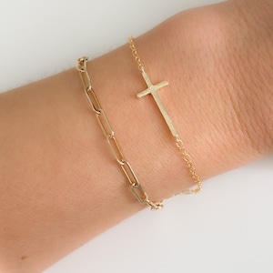 May include: Two gold bracelets on a wrist. One bracelet is a simple chain with a cross pendant. The other bracelet is a thicker chain with larger links.