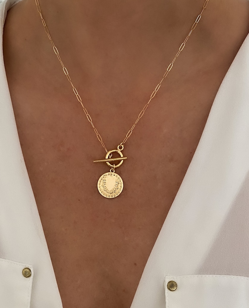 K&ouml;nnte beinhalten: Eine goldene Kettenkette mit einem Toggle-Verschluss und einem runden Anh&auml;nger mit einem Lorbeerkranz-Design.