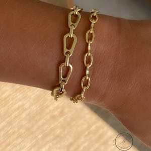 May include: Two gold-colored chain bracelets. One bracelet features a series of elongated, open links, while the other has a more traditional oval link design. The bracelets are displayed on a person's wrist, showcasing their design and style.