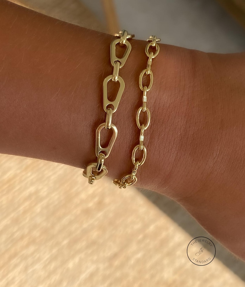 May include: Two gold-colored chain bracelets. One bracelet features a series of elongated, open links, while the other has a more traditional oval link design. The bracelets are displayed on a person's wrist, showcasing their design and style.