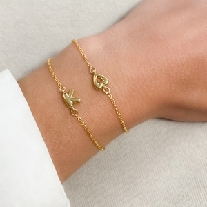 May include: Two gold chain bracelets with a heart charm and a letter K charm. The bracelets are on a person's wrist.