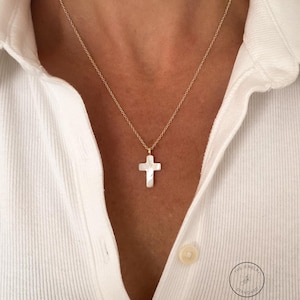 May include: A gold-colored chain necklace with a white cross pendant. The cross pendant has a pearlescent finish. The necklace is worn over a white collared shirt. The Jewelry Standard logo is visible on the shirt.