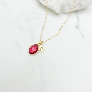 May include: A gold chain necklace with a small, red gemstone pendant and a gold charm with a letter 'A' on it.