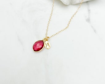May include: A gold chain necklace with a small, red gemstone pendant and a gold charm with a letter 'A' on it.