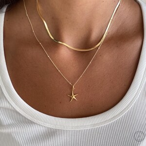 May include: A gold-colored necklace set featuring two necklaces. The top necklace has a flat, wavy design, while the bottom necklace has a small starfish pendant. The necklaces are worn on a person wearing a white ribbed tank top.