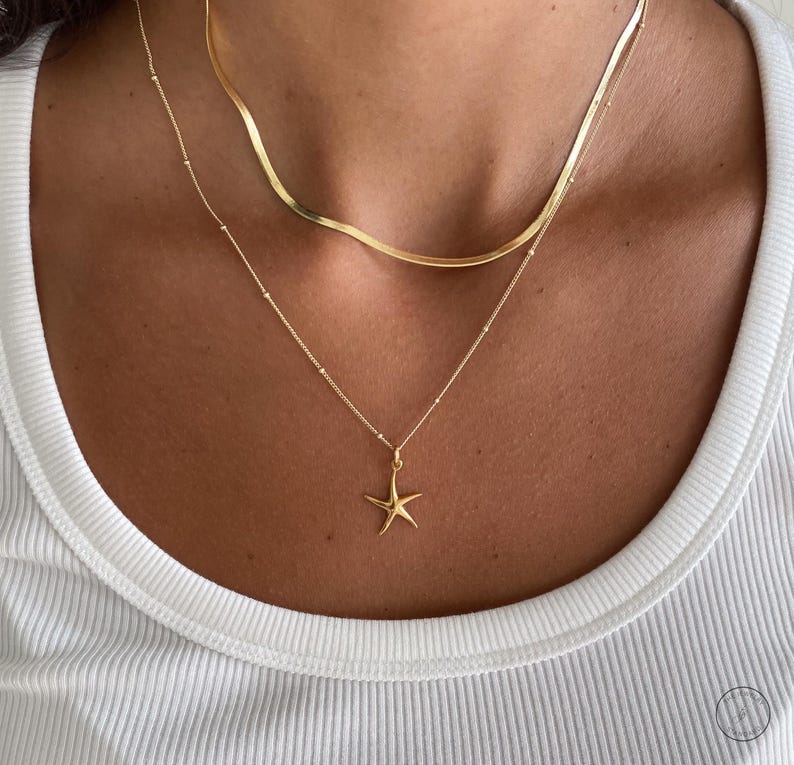May include: A gold-colored necklace set featuring two necklaces. The top necklace has a flat, wavy design, while the bottom necklace has a small starfish pendant. The necklaces are worn on a person wearing a white ribbed tank top.