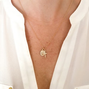May include: A gold-colored necklace with two pendants. One pendant is a small cross, and the other is a circular medallion. The necklace is worn against a white blouse.