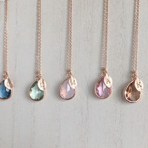 May include: Five rose gold necklaces with teardrop-shaped pendants in various colors: blue, green, pink, and amber. Each pendant has a small leaf charm and a single initial. The necklaces hang on a white wooden surface.