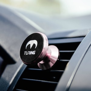 Pu&ograve; includere: Un supporto magnetico per telefono da auto in oro rosa con una base circolare nera. La base ha la scritta "TURING" stampata in lettere bianche e un logo bianco.