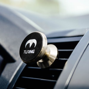Pu&ograve; includere: Un supporto magnetico per telefono cellulare da auto in argento e nero con la scritta "TURING" stampata sulla parte nera. Il supporto &egrave; fissato a una bocchetta di ventilazione dell'auto.