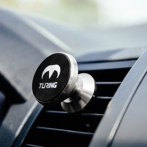 Pu&ograve; includere: Un supporto magnetico per telefono cellulare da auto nero e argento con la scritta "TURING" stampata sul davanti. Il supporto &egrave; fissato a una bocchetta di ventilazione dell'auto.