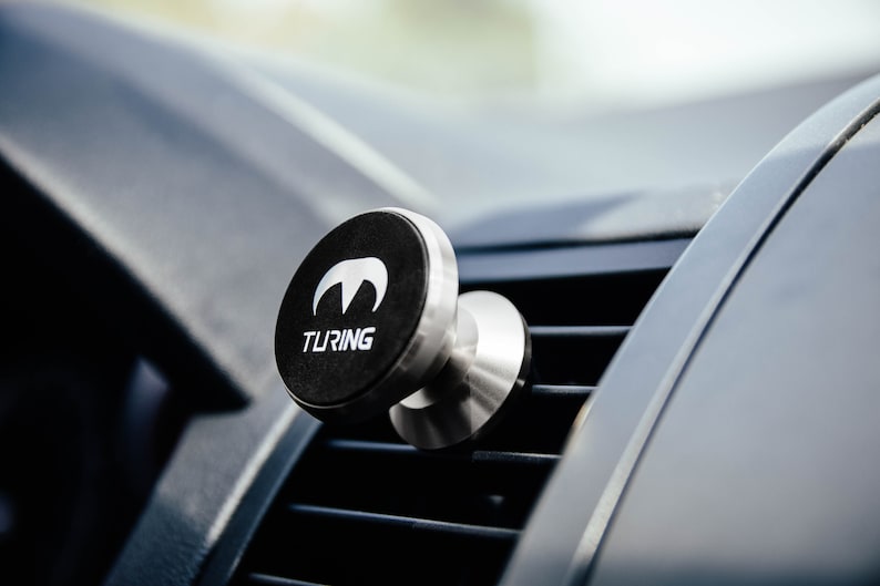 Pu&ograve; includere: Un supporto magnetico per telefono cellulare da auto nero e argento con la scritta "TURING" stampata sul davanti. Il supporto &egrave; fissato a una bocchetta di ventilazione dell'auto.