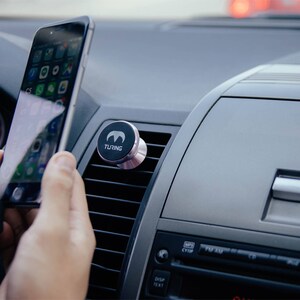 Pu&ograve; includere: Un supporto magnetico nero per telefono con la scritta "TURING" &egrave; attaccato alla bocchetta di aerazione di un'auto. Una persona tiene uno smartphone in mano.