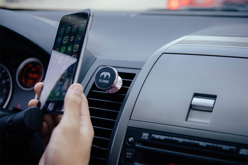 Pu&ograve; includere: Un supporto magnetico nero per telefono con la scritta "TURING" &egrave; attaccato alla bocchetta di aerazione di un'auto. Una persona tiene uno smartphone in mano.