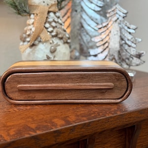 Puede incluir: Una caja de madera con un cajón, hecha de madera de nogal y arce. La caja tiene una forma redondeada y un acabado liso.