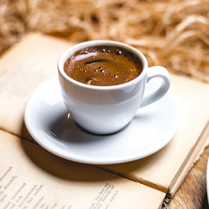 May include: A white ceramic coffee cup filled with dark coffee, resting on a matching saucer. The cup and saucer are placed on an open book with aged pages. The background features a blurred, textured surface.