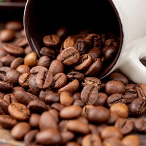 K&ouml;nnte beinhalten: Eine wei&szlig;e Kaffeetasse mit braunem Innenraum ist umgekippt und versch&uuml;ttet ger&ouml;stete Kaffeebohnen auf eine Holzoberfl&auml;che. Die Bohnen sind in verschiedenen Braunt&ouml;nen gehalten und haben ein reichhaltiges, strukturiertes Aussehen. Kaffee-Thema.