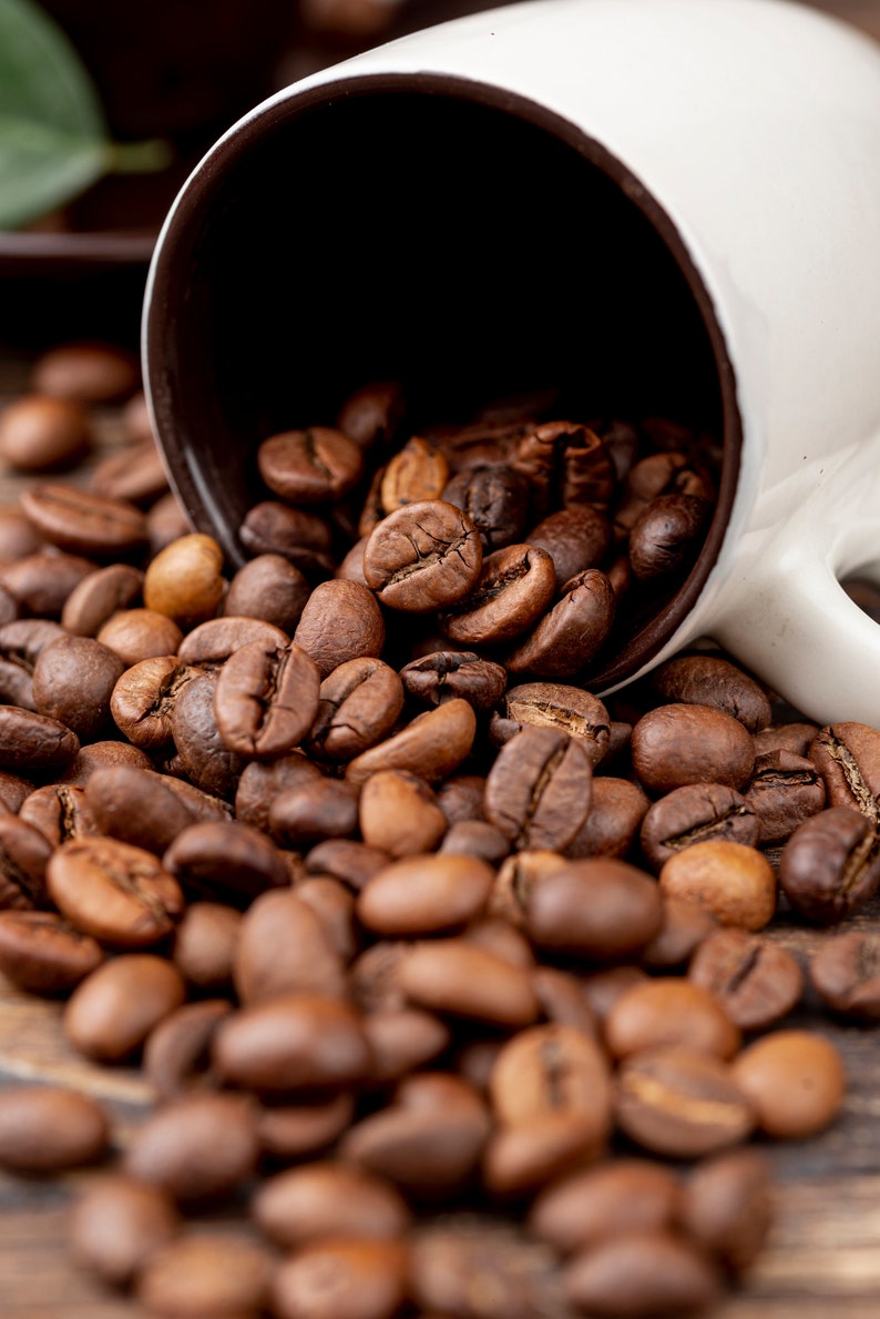 K&ouml;nnte beinhalten: Eine wei&szlig;e Kaffeetasse mit braunem Innenraum ist umgekippt und versch&uuml;ttet ger&ouml;stete Kaffeebohnen auf eine Holzoberfl&auml;che. Die Bohnen sind in verschiedenen Braunt&ouml;nen gehalten und haben ein reichhaltiges, strukturiertes Aussehen. Kaffee-Thema.