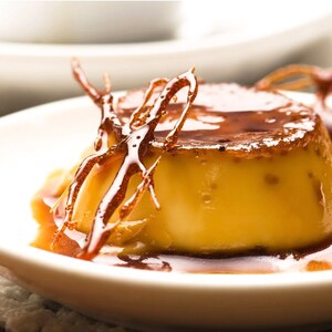May include: A close-up of a creamy yellow flan dessert topped with a caramel sauce and a decorative caramel swirl. The flan is on a white plate.