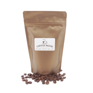 K&ouml;nnte beinhalten: Ein brauner Papiersack mit Kaffeebohnen und einem wei&szlig;en Etikett mit der Aufschrift "Coffee Nook, Roasted in the Sea, Micro Roasting Co." Ger&ouml;stete Kaffeebohnen sind am Boden des Beutels verstreut.