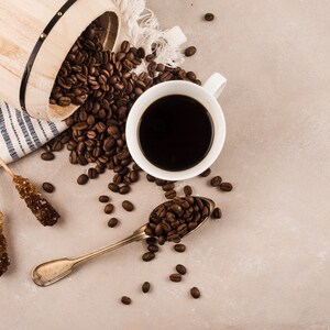 K&ouml;nnte beinhalten: Draufsicht auf eine Kaffeekomposition. Eine wei&szlig;e Tasse mit dunklem Kaffee steht neben einem L&ouml;ffel mit Kaffeebohnen. Kaffeebohnen sind um einen Holzeimer und zwei Kandiszucker herum verstreut.