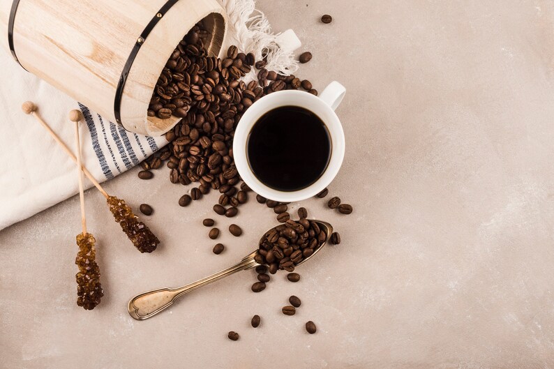 K&ouml;nnte beinhalten: Draufsicht auf eine Kaffeekomposition. Eine wei&szlig;e Tasse mit dunklem Kaffee steht neben einem L&ouml;ffel mit Kaffeebohnen. Kaffeebohnen sind um einen Holzeimer und zwei Kandiszucker herum verstreut.