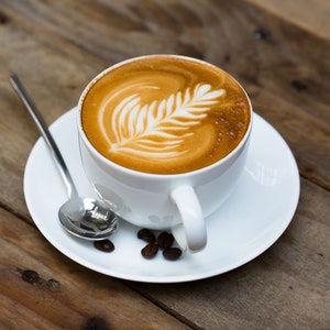 K&ouml;nnte beinhalten: Eine wei&szlig;e Kaffeetasse gef&uuml;llt mit einem Latte, mit einem Blattmuster im Schaum. Die Tasse steht auf einer passenden Untertasse, mit einem silbernen L&ouml;ffel und ein paar Kaffeebohnen auf einer Holzoberfl&auml;che.