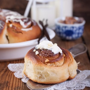 May include: A close-up of a cinnamon roll topped with a swirl of white frosting and chopped nuts. The roll is sitting on a silver serving spoon and a white doily.