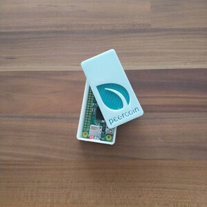 Puede incluir: Una caja de plástico blanca con un logotipo de hoja verde y el texto "peercoin" en la tapa. La caja está abierta y se puede ver un pequeño circuito verde en el interior.