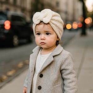 May include: A young child wearing a cream-colored knitted turban with a bow detail. The child is also wearing a light gray coat with black buttons and a white dress. The background shows a blurred city street.