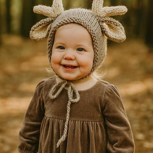 May include: A smiling toddler wearing a knitted beige deer antler hat and a brown velvet dress. The hat has two large, upright antlers and ear-shaped flaps. The dress has long sleeves and a tied neckline.