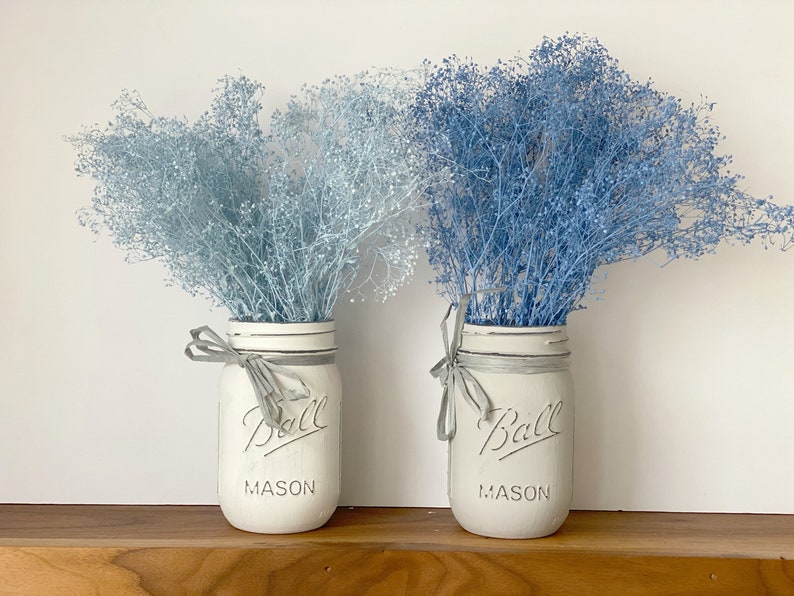 May include: Two white mason jars with gray lids and gray ribbon bows, filled with blue and light blue dried baby's breath.