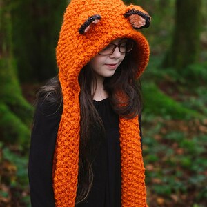 May include: An orange crocheted fox hat and scarf set. The hood features fox ears with black and tan accents. The scarf is long and has pockets. The set is made of a chunky, textured yarn.
