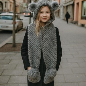 May include: A child wearing a gray knitted hat with bear ears and a matching scarf with mittens. The hat and scarf set are made of a chunky knit material. The child is wearing a black coat.