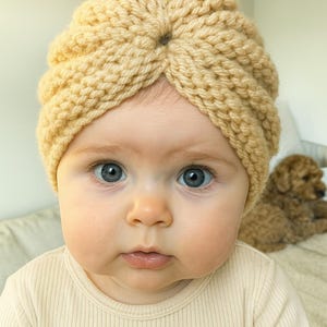 May include: A baby wearing a light tan knitted turban hat. The hat is made of a chunky yarn and has a central knot detail. The baby has blue eyes and is wearing a light tan long-sleeved top.