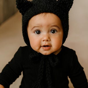May include: A baby wearing a black knitted hat with cat ears and a matching black outfit. The hat has a tie under the chin with two pom-poms. The baby is looking directly at the camera.