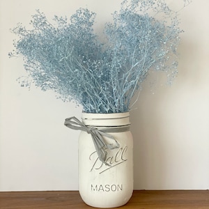 May include: A white painted mason jar with a gray ribbon and the text "Ball Mason" on the front. The jar is filled with a bouquet of light blue baby's breath.