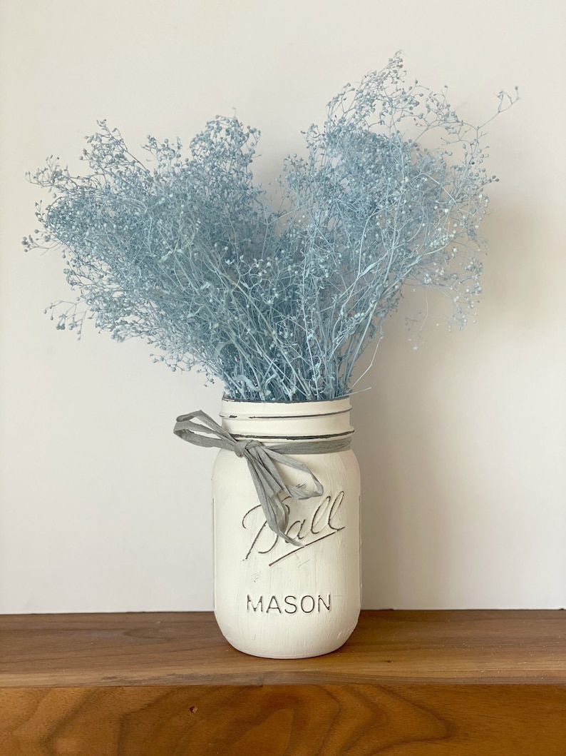 May include: A white painted mason jar with a gray ribbon and the text "Ball Mason" on the front. The jar is filled with a bouquet of light blue baby's breath.