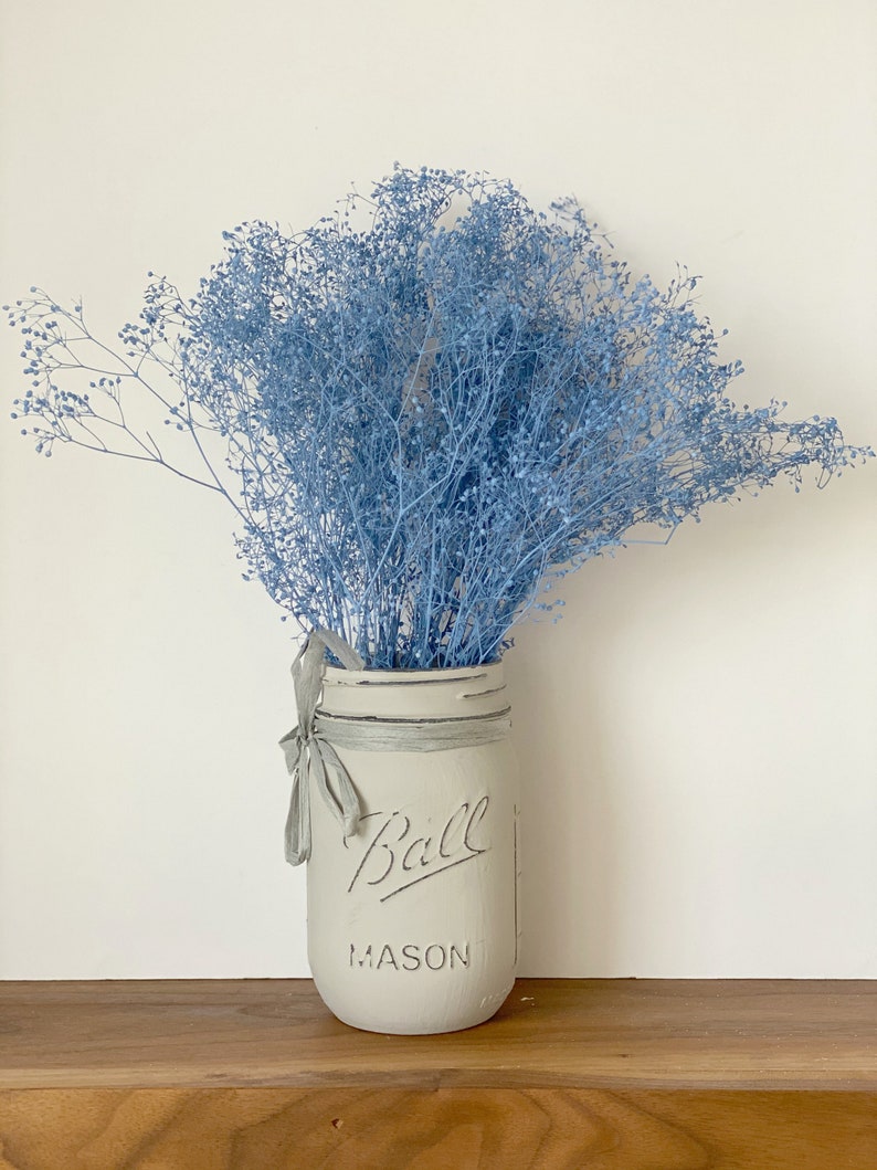 May include: A light blue bouquet of baby's breath flowers in a white painted Ball Mason jar with a gray ribbon.