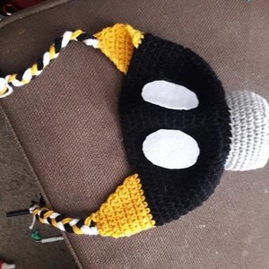 May include: A black, yellow, and gray crocheted hat with white felt eyes. The hat has a long, braided tail.