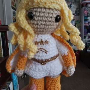 May include: A handmade crochet doll with a light brown face, large black eyes, and curly yellow hair. The doll wears a white dress with gold accents and orange and white wings. The doll's legs and boots are orange.