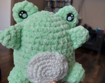 Könnte beinhalten: Ein grünes, gehäkeltes Frosch-Plüschtier mit weißen Häkeldetails. Der Frosch hat schwarze Augen mit weißen Pupillen.
