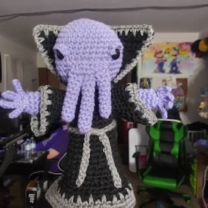 Puede incluir: Muñeca de ganchillo hecha a mano con una cabeza morada y tentáculos, ojos negros y una túnica negra con ribetes grises. La muñeca tiene los brazos extendidos y un cinturón gris. Un artículo único y hecho a mano.