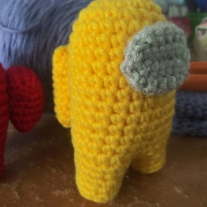 May include: A yellow crocheted plush toy shaped like the character from the video game Among Us. The toy has a gray crocheted visor.