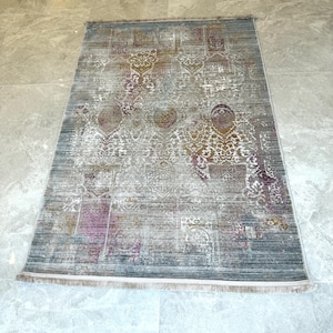 May include: Rectangular rug with a distressed, vintage appearance. The rug has a grey and white background with ornate floral and geometric patterns in purple and gold. It has a fringed edge.