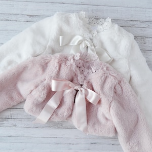 Op de afbeelding: Twee nep bontjasjes, een wit en een roze, met satijnen strikjes en kantversiering. De jasjes liggen op een wit houten oppervlak.